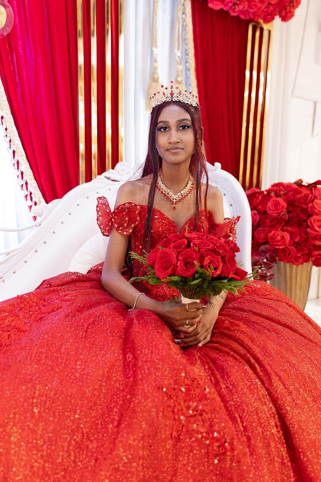 Quinceañera in a red ball gown seated on a white throne with roses